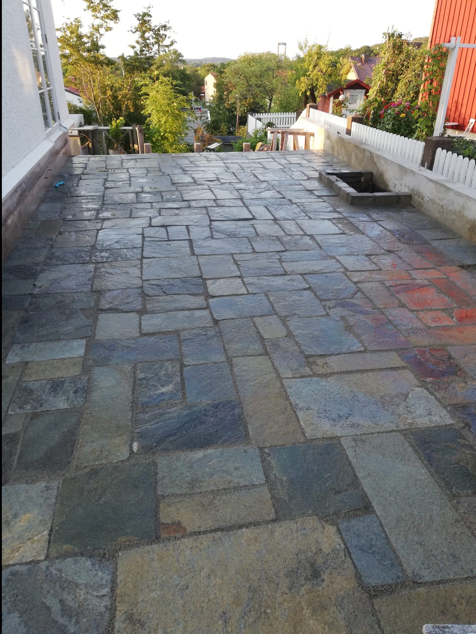 Kamienny taras z nieregularnych płyt w odcieniach szarości i błękitu, z widokiem na zieleń ogrodu i elementy architektury w tle. Widoczne detale wykończenia.