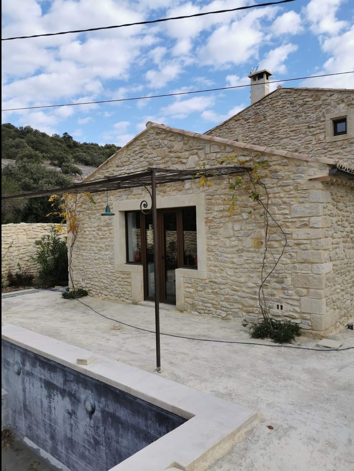 Kamienny dom z pergolą i częściowo widocznym basenem. Widok na elewację, okna z ciemnymi ramami, zielone pnącze na pergoli. Błękitne niebo z chmurami w tle.