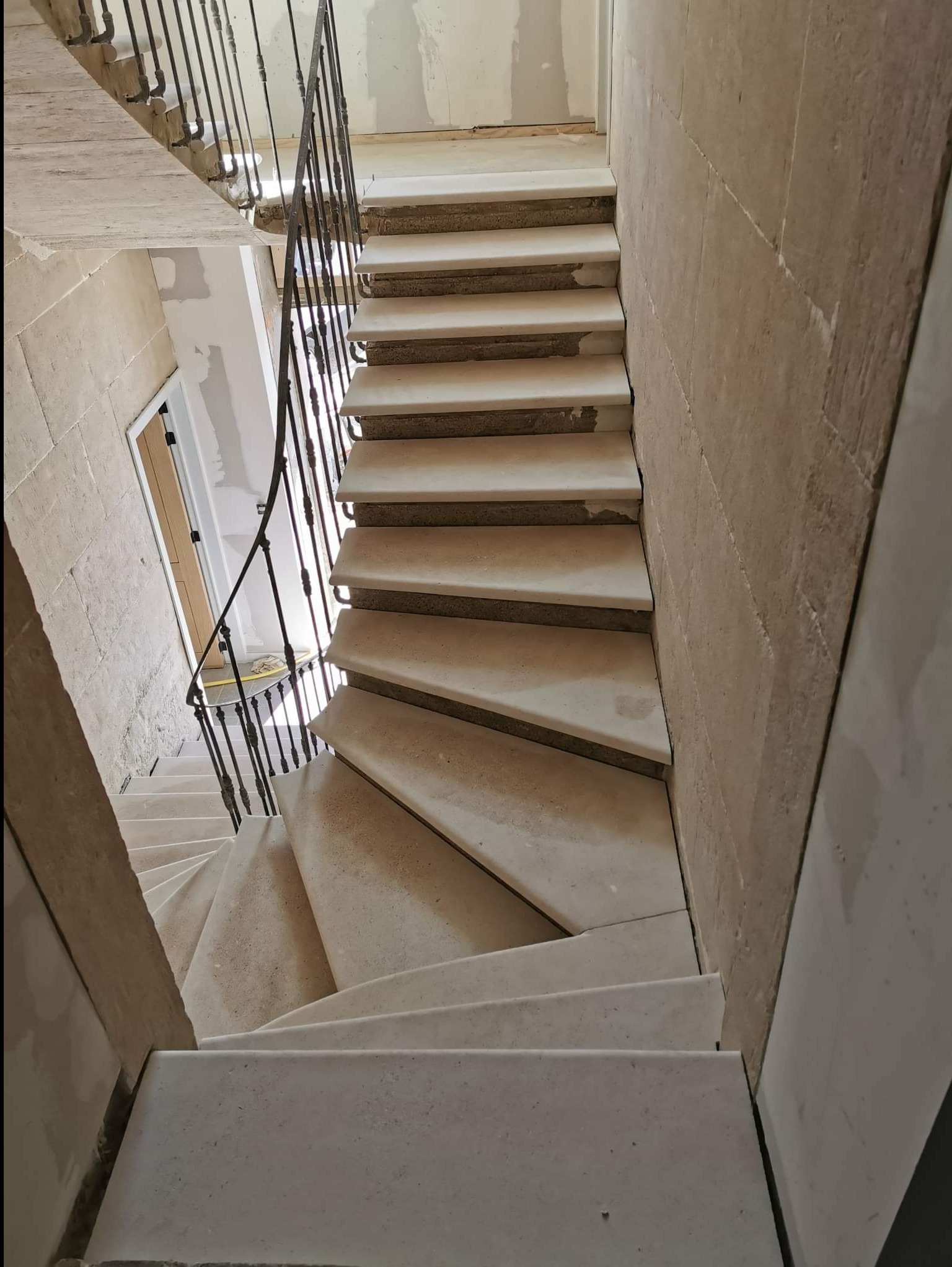 Wewnętrzne schody kamienne z kutą balustradą w trakcie montażu, widok z góry. Surowe wykończenie ścian, brak listew przypodłogowych, etap prac wykończeniowych.