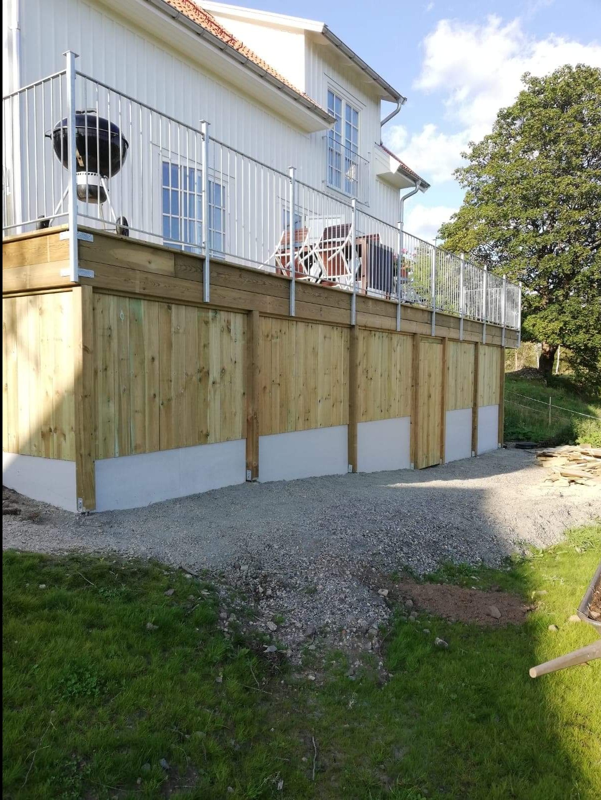 Wykończony taras drewniany przy domu z metalową balustradą. Widoczne deski tarasowe, podkonstrukcja z drewna i betonowe elementy wsporcze. Na tarasie stoi grill.