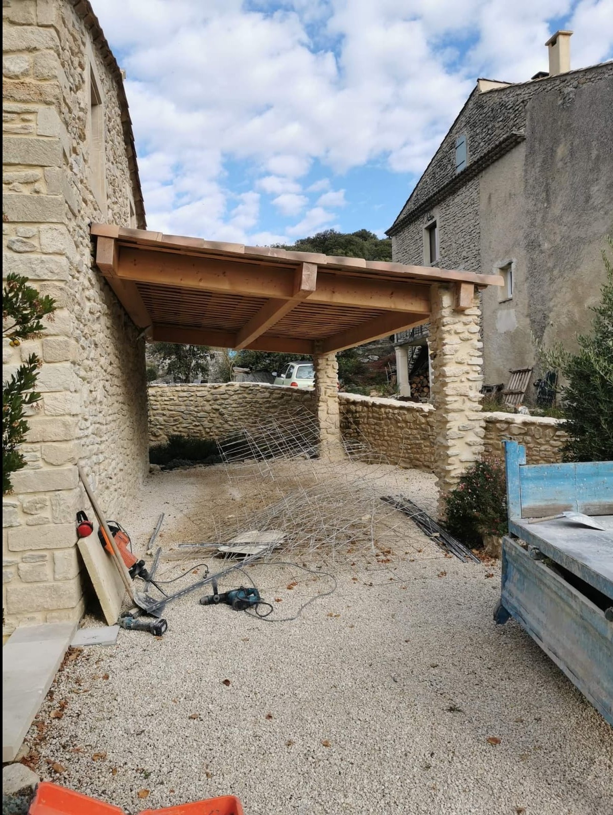 Drewniana pergola z kamiennymi kolumnami przy kamiennym domu w trakcie montażu, widoczne narzędzia i materiały budowlane na żwirowym podłożu.