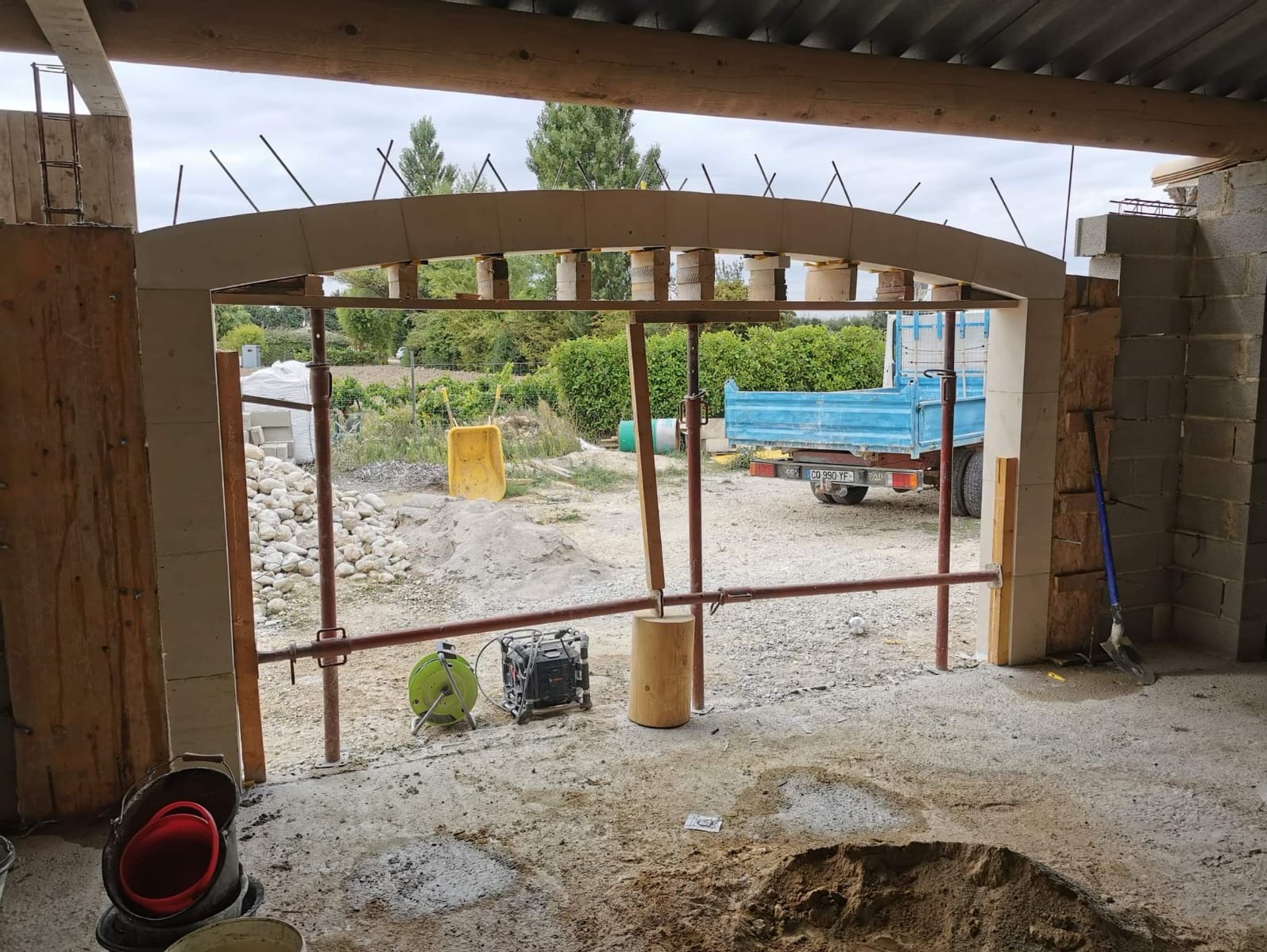 Wnętrze budynku w trakcie budowy, widoczny łuk z betonu komórkowego wsparty stemplami. Na zewnątrz teren budowy, piasek, gruz i samochód ciężarowy.