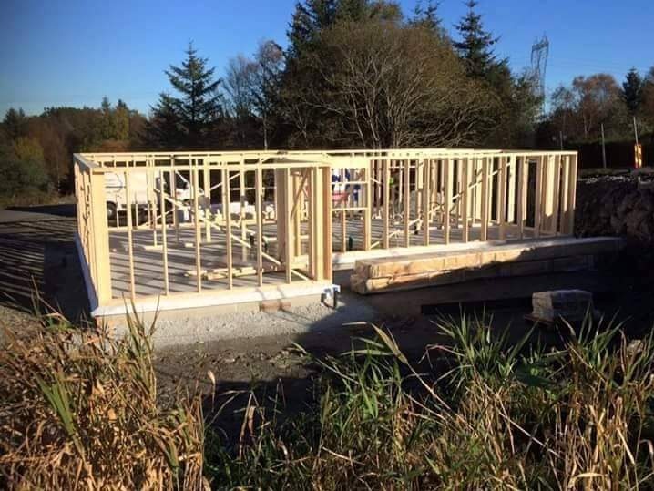 Konstrukcja szkieletowa domu w trakcie budowy, widok z zewnątrz na drewniany szkielet, zieleń w tle, słoneczny dzień.