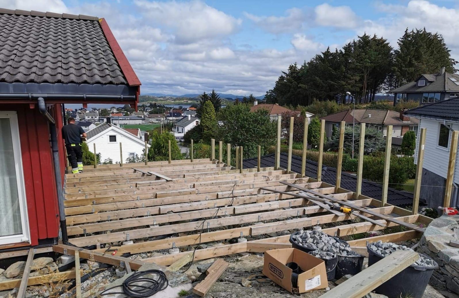 Montaż drewnianego tarasu: szkielet z belek, pionowe słupki, widok na krajobraz miejski. Prace w toku, materiały budowlane, lekko pochmurne niebo.