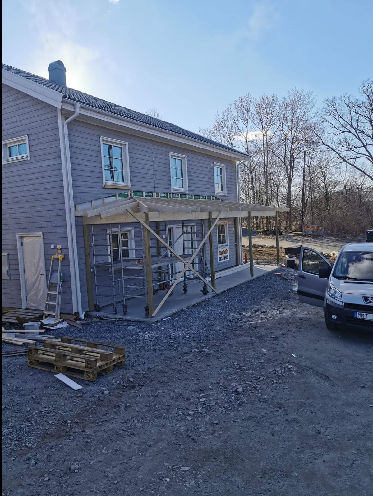 Szary dom z drewnianą konstrukcją zadaszenia tarasu w trakcie budowy. Widoczne rusztowania, paleta i gruz. Samochód dostawczy obok.