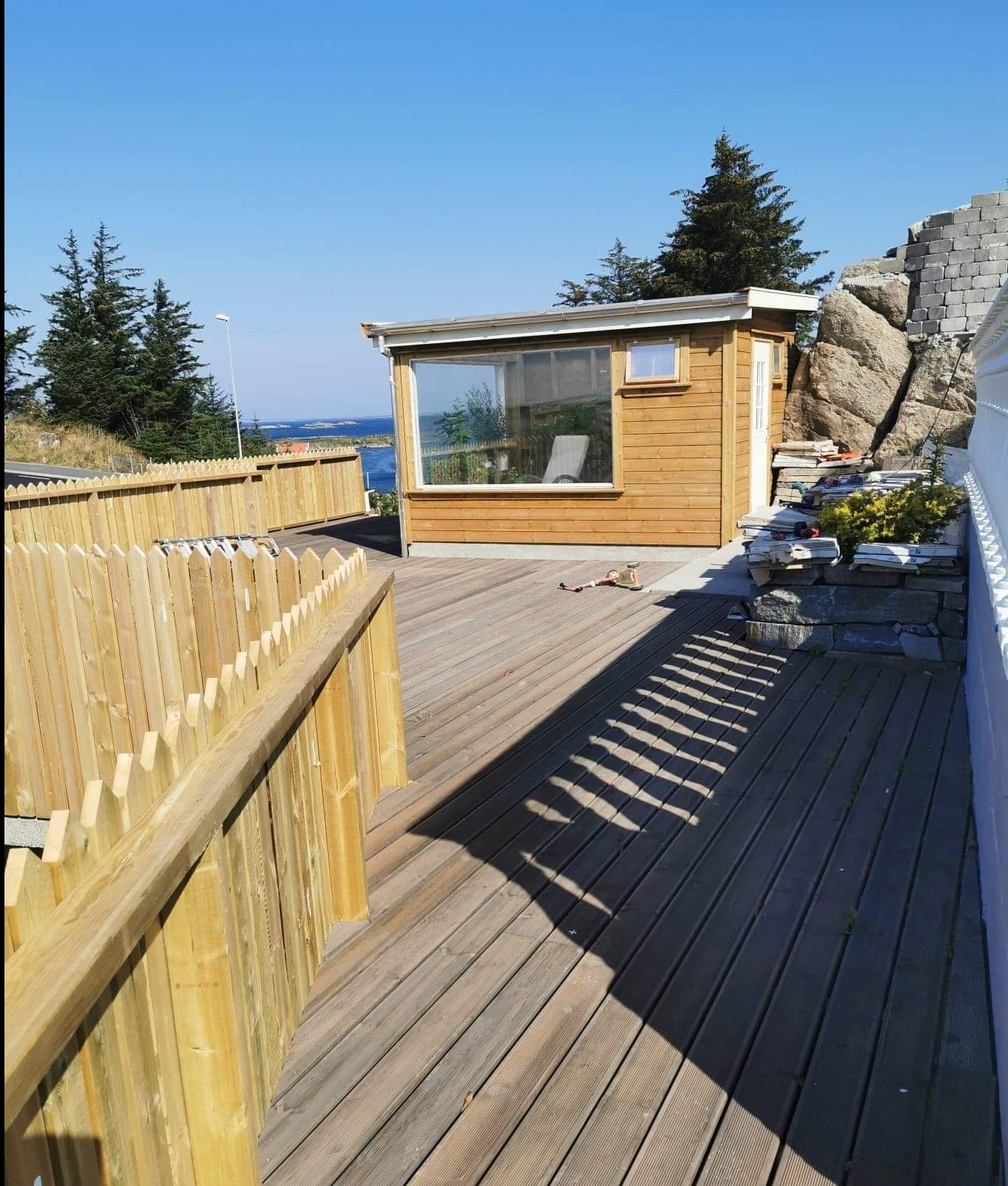 Drewniany taras z jasnym ogrodzeniem i domkiem letniskowym z dużym oknem na tle błękitnego nieba i morza, w słoneczny dzień.