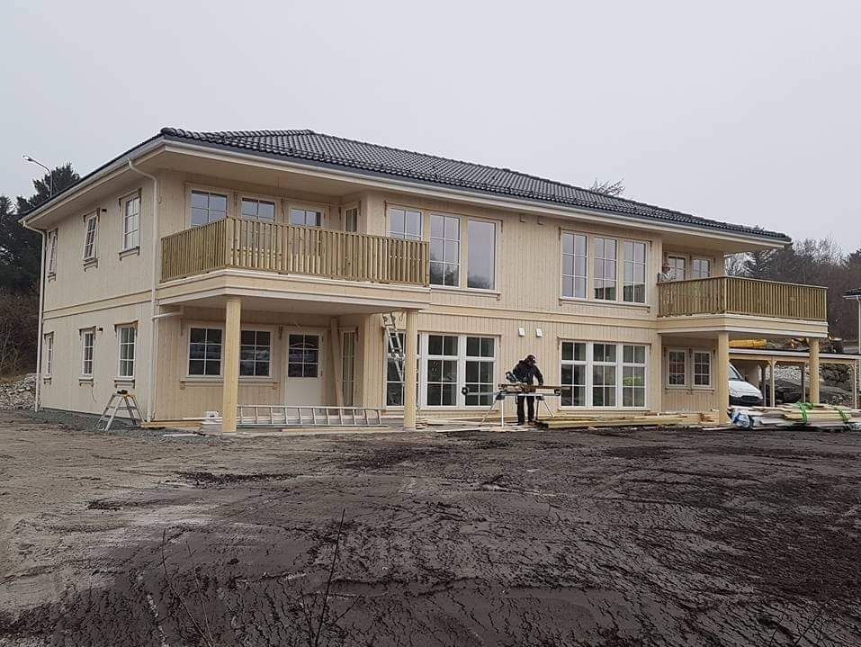 Dwukondygnacyjny dom szkieletowy z balkonami, w trakcie budowy. Widoczny stolarz przy pracy, surowe drewno i teren budowy. Dom z ciemną dachówką.
