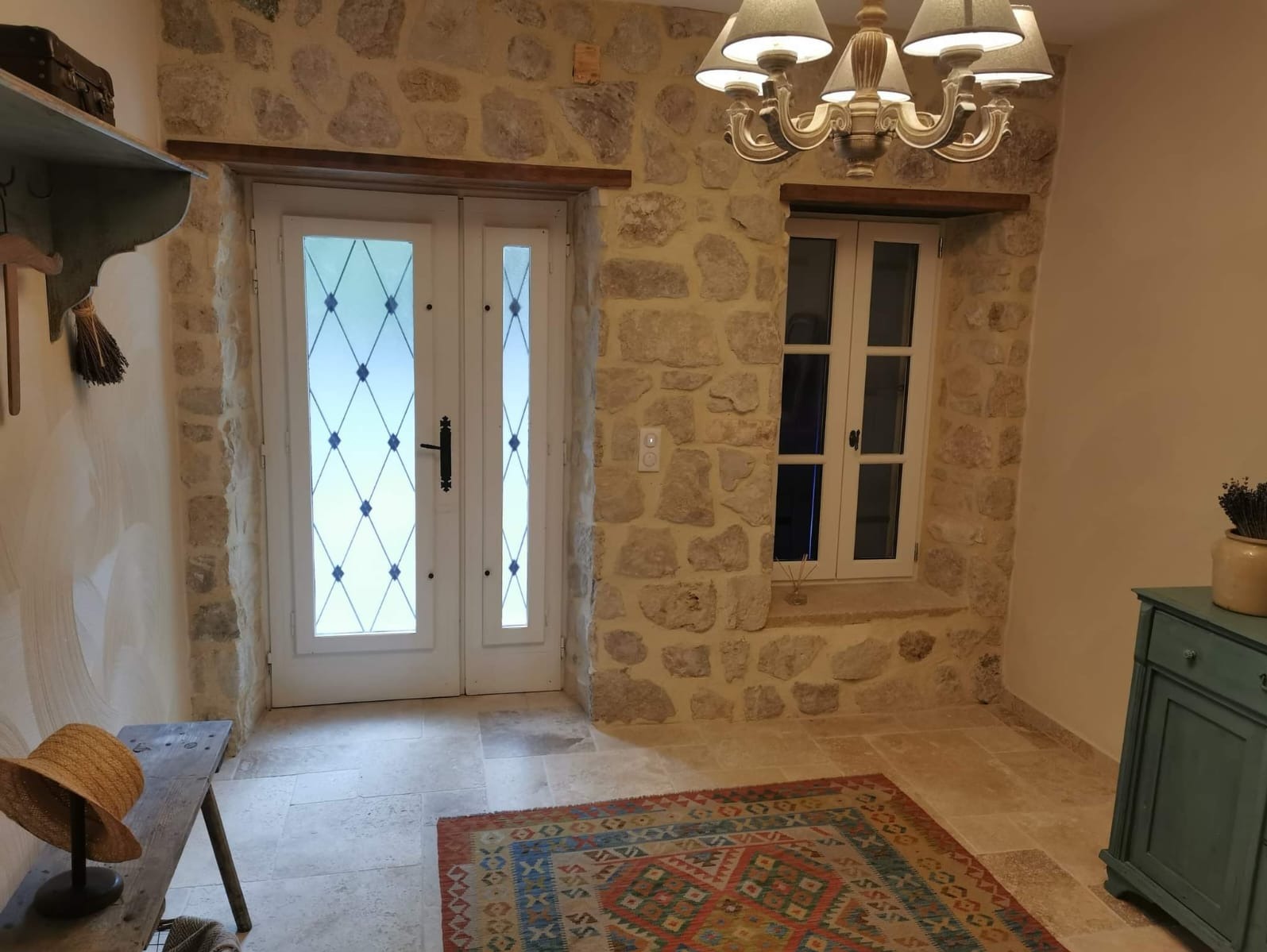 Kamienne ściany z oknem i przeszklonymi drzwiami. Wnętrze z dywanem, lampą i drewnianą ławką z kapeluszem. Widoczne elementy dekoracyjne.