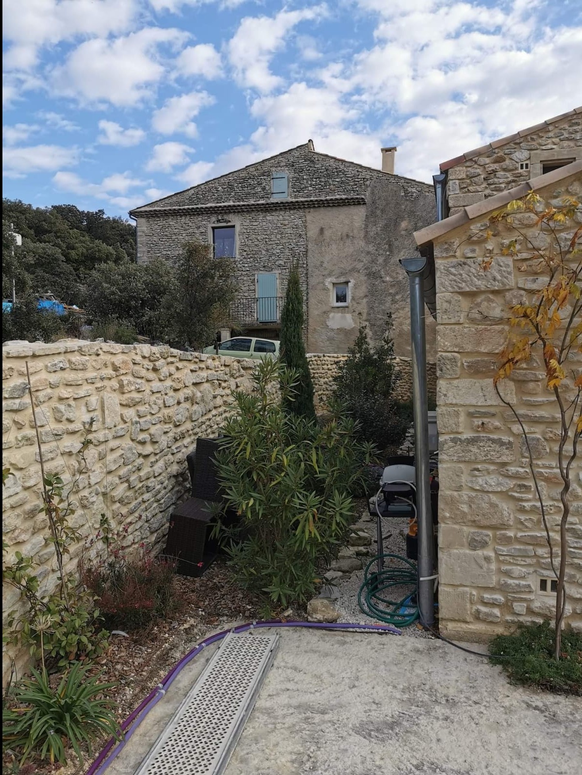 Kamienna zabudowa domu z tarasem i murem, zieleń ozdobna, błękitne niebo. Widoczny fragment tarasu z metalową kratką odpływową.