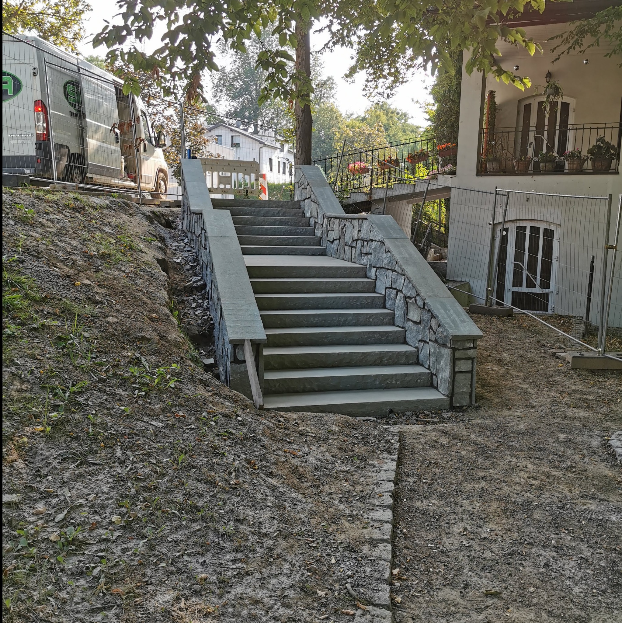 Kamienne schody zewnętrzne z balustradami, prowadzące do domu z balkonem. Widoczny fragment ogrodu i samochód dostawczy w tle. Dzień słoneczny.