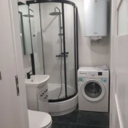 Niewielka łazienka z kabiną prysznicową z czarnymi profilami, umywalką z szafką, toaletą, pralką i bojlerem elektrycznym na ścianie, wykończona w bieli i czerni.