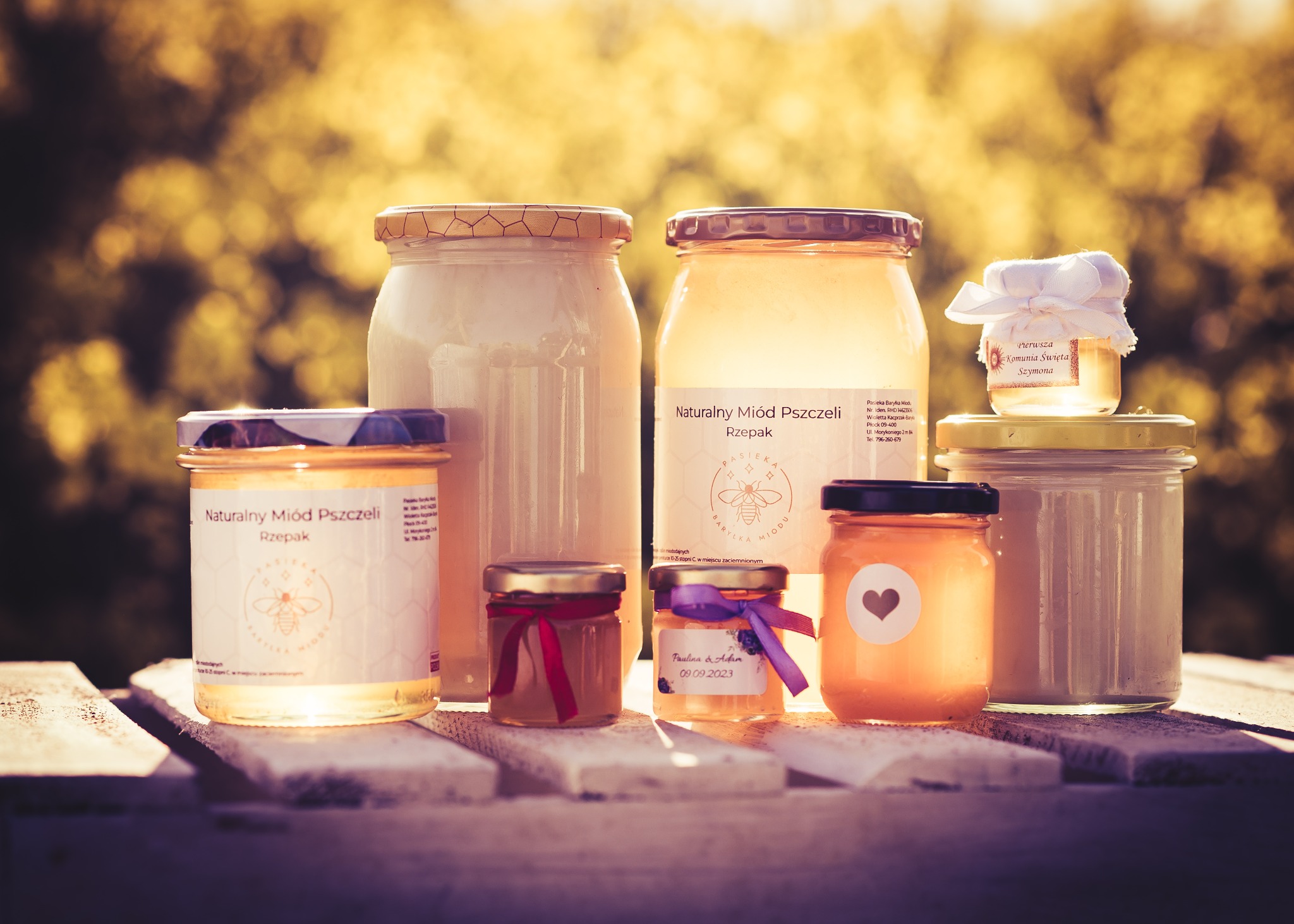 Zestaw słoików z miodem różnego rodzaju i wielkości, niektóre ozdobione wstążkami i etykietami okolicznościowymi, ustawione na drewnianym podeście na tle rozmytej zieleni.