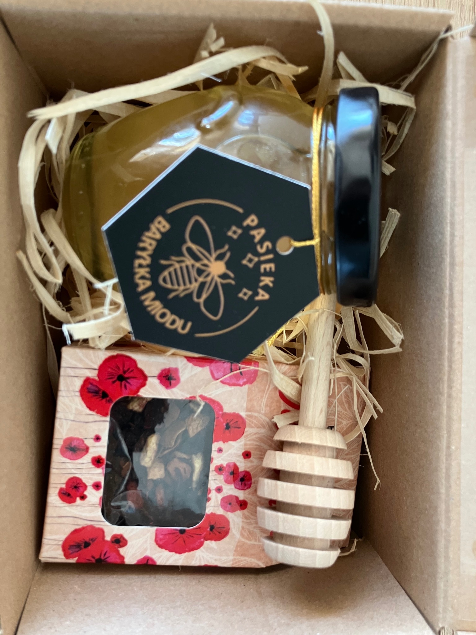 Kosz prezentowy: słoik miodu z czarną etykietą 'Pasieka Baryczka Miodu', nabierak i pudełko herbaty w kartonowym pudełku z wypełnieniem.