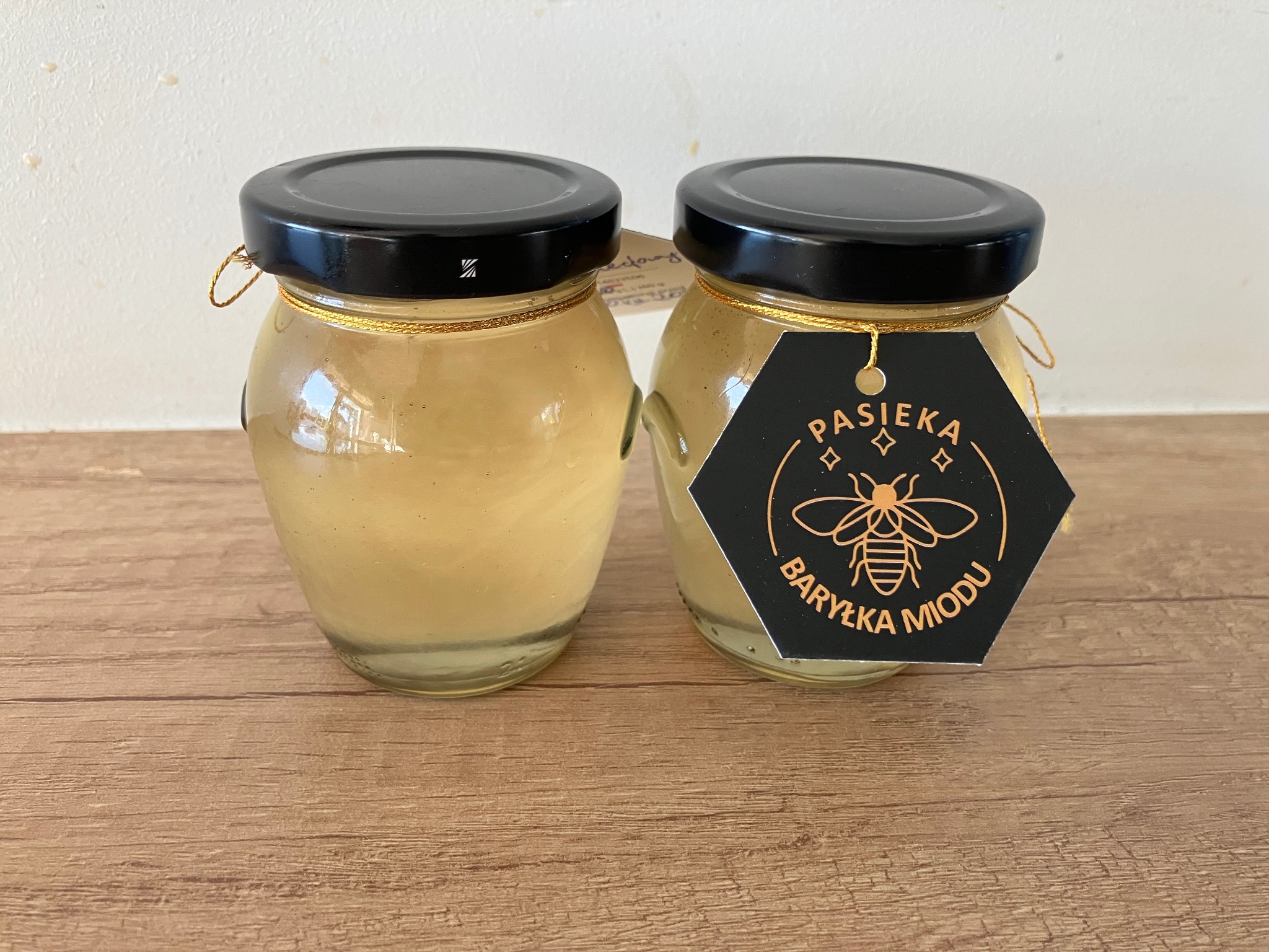Dwa słoiki z miodem na drewnianym blacie, jeden z czarną etykietą w kształcie plastra miodu z napisem 'Pasieka Baryłka Miodu'. Słoiki ozdobione złotym sznurkiem.