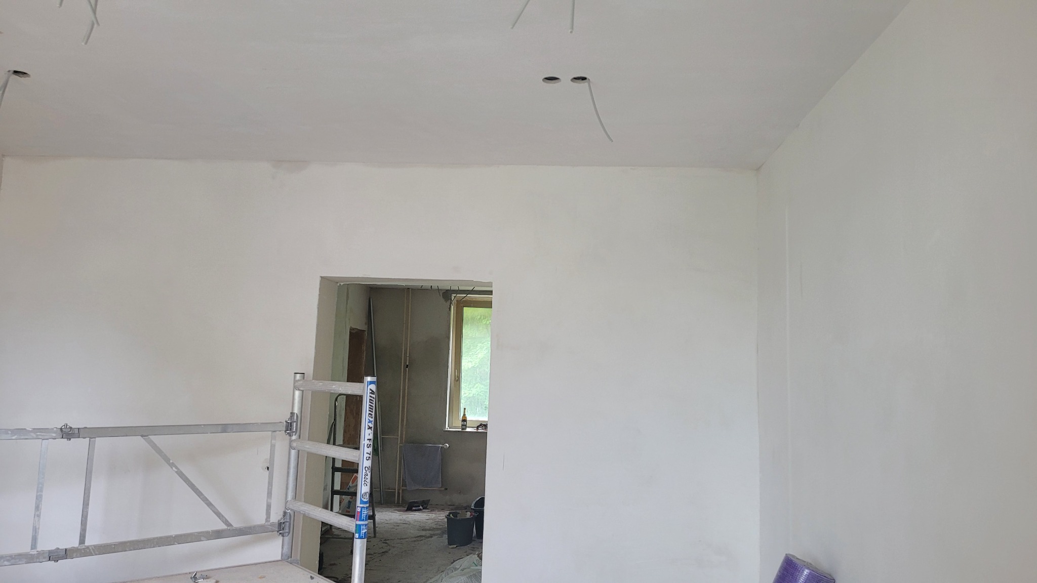 Pokój w trakcie przygotowania do malowania, świeżo wyszpachlowane ściany i sufit, widoczne kable elektryczne, aluminiowy rusztowanie w rogu pomieszczenia, okno w tle.