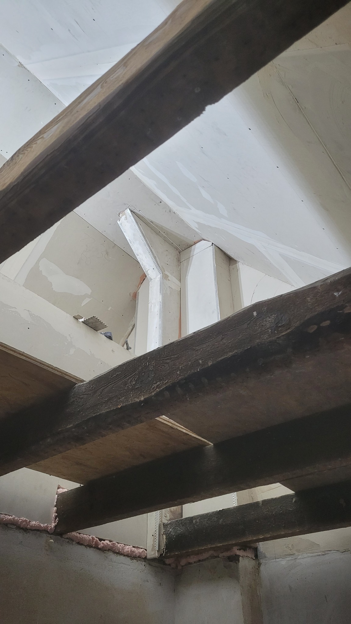 Sufit poddasza w trakcie prac wykończeniowych, widoczne drewniane belki konstrukcyjne, płyty gipsowo-kartonowe z nałożoną siatką wzmacniającą i częściowo wyszpachlowane, różowa pianka izolacyjna...