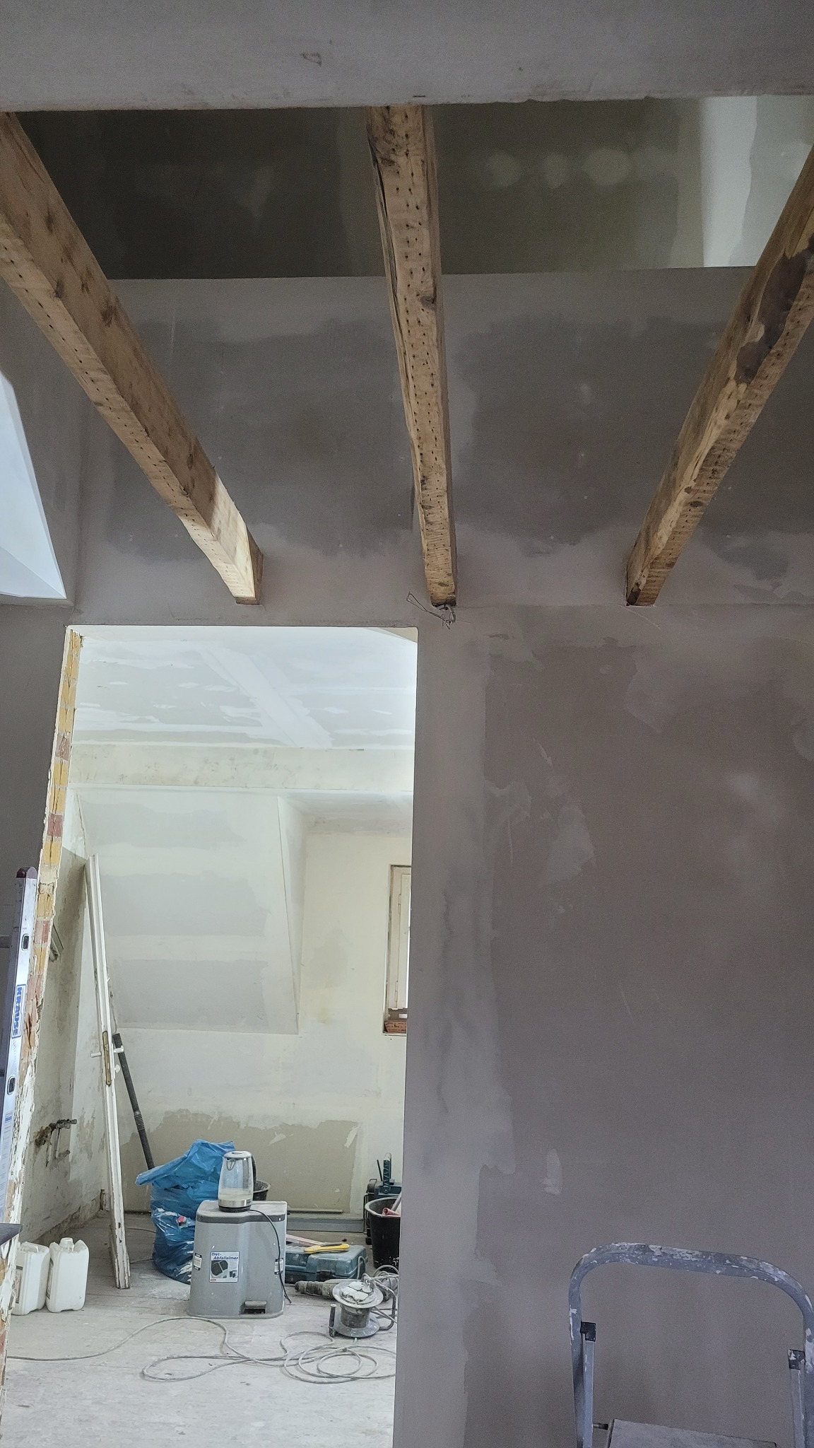 Wnętrze w trakcie remontu: surowa ściana po szpachlowaniu, widoczne drewniane belki stropowe i sprzęt budowlany w pomieszczeniu obok.
