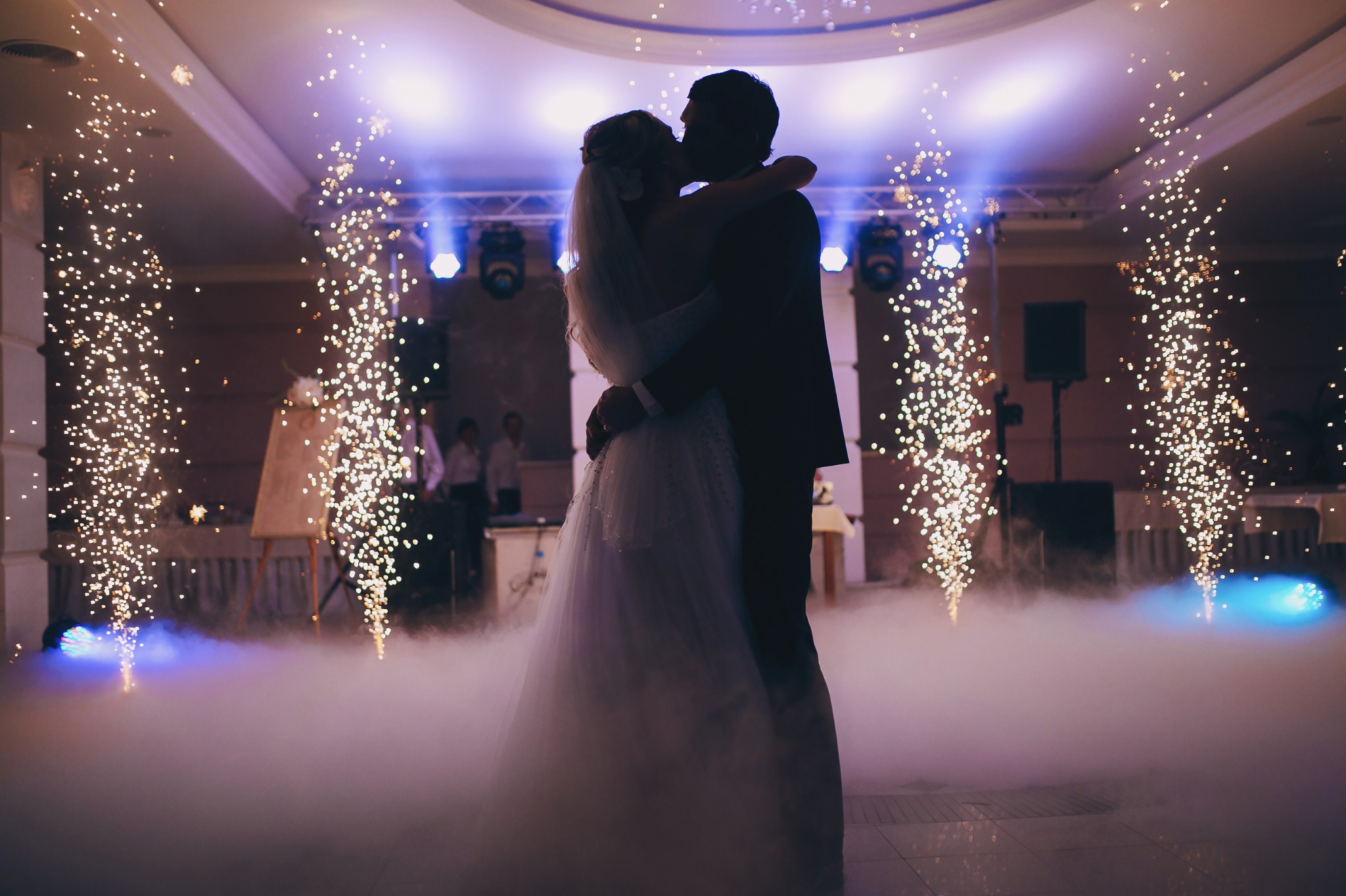 Silhouettes of a bride and groom kissing amidst a smoky dance floor illuminated by spotlights and sparkling pyrotechnics during a wedding reception.
