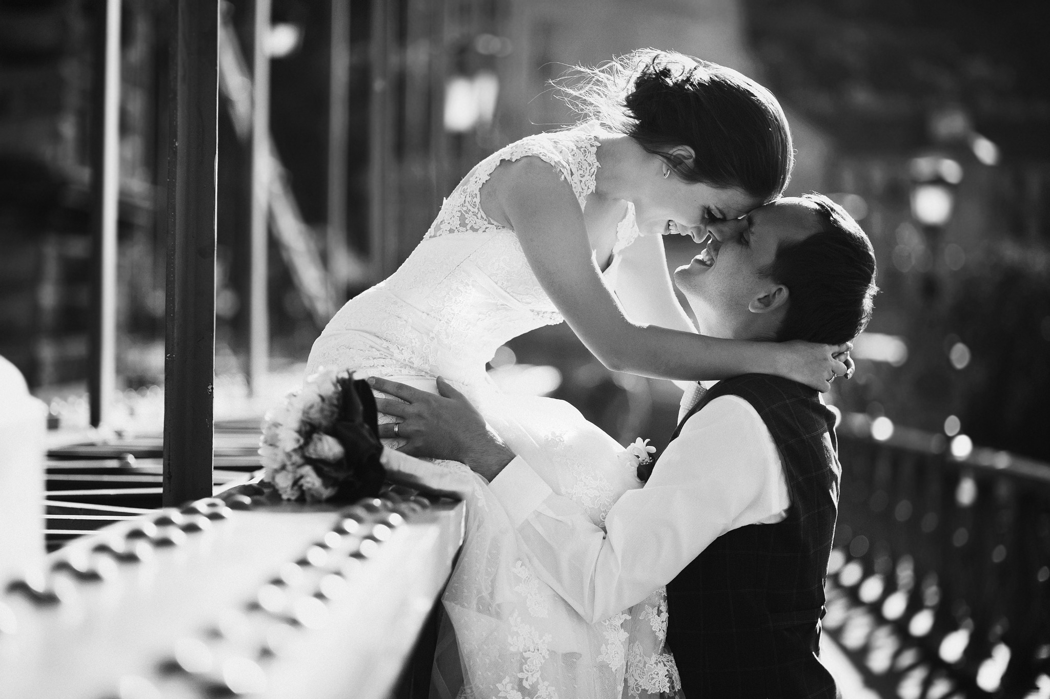 Czarno-białe zdjęcie uśmiechniętej panny młodej w koronkowej sukni, siedzącej na barierce i obejmującej pana młodego w kamizelce w kratę, z bukietem ślubnym obok, w tle rozmyte światła.