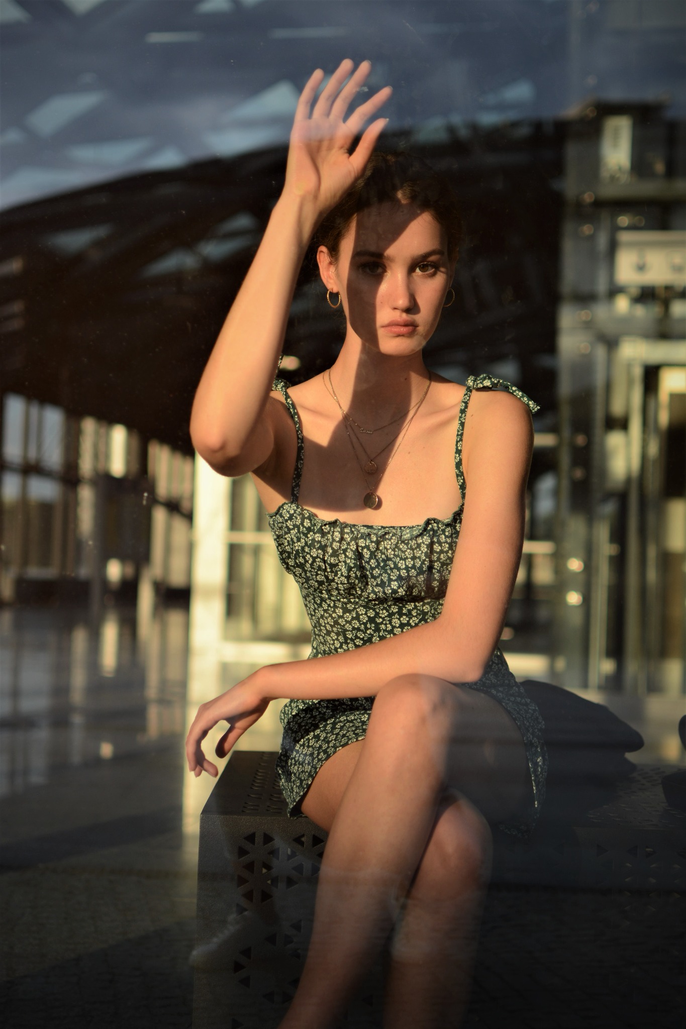 Młoda kobieta w letniej sukience siedzi na metalowej ławce w przeszklonym wnętrzu, z dłonią uniesioną do szyby, odbijając światło słoneczne.