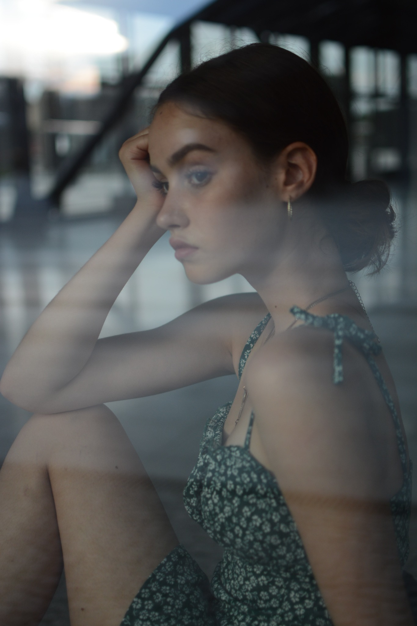 Zamyślona młoda kobieta w kwiecistej sukience, oparta o szybę z odbiciem miejskiego krajobrazu, portret z profilu.