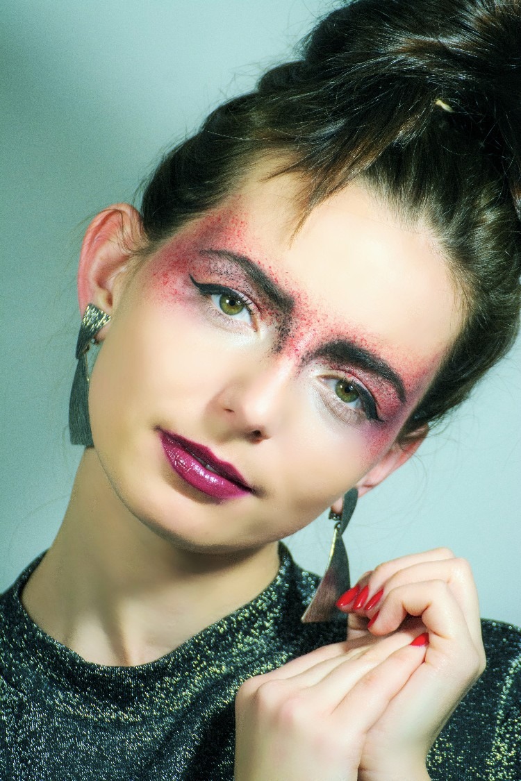 Portret kobiety z artystycznym makijażem oczu w odcieniach czerwieni i czerni, ciemnofioletowe usta, geometryczne kolczyki i czerwone paznokcie.