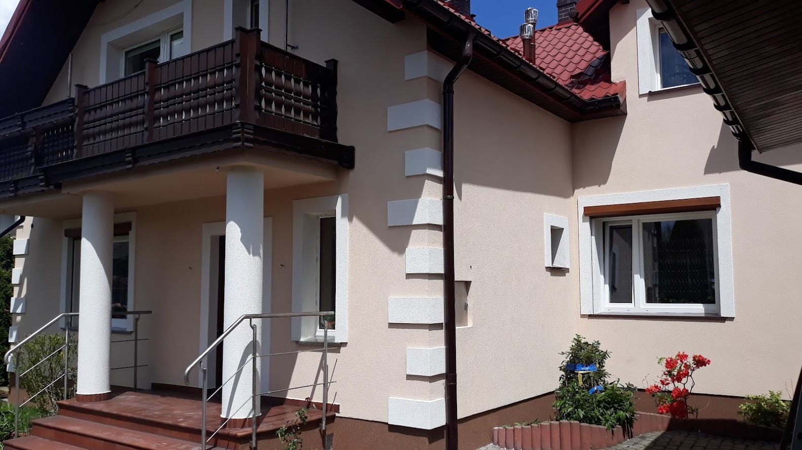 Jasny, dwukondygnacyjny dom z beżową elewacją, białymi detalami architektonicznymi wokół okien i na narożnikach, ciemnobrązowym balkonem i czerwonym dachem, sfotografowany w słoneczny dzień.
