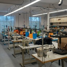 ArTy Szwalnia - Jasne wnętrze szwalni z maszynami do szycia, materiałami i akcesoriami. Widoczne stanowiska pracy, regał z pudełkami i okna w tle.