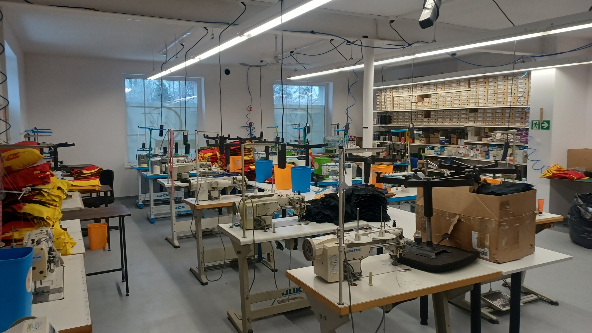 Jasne wnętrze szwalni z maszynami do szycia, materiałami i akcesoriami. Widoczne stanowiska pracy, regał z pudełkami i okna w tle.