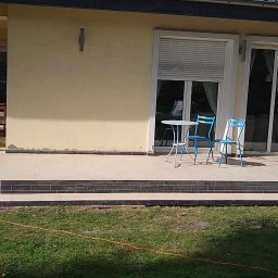 ARR-BUD - Żółta elewacja domu z tarasem wyłożonym płytkami, na którym stoi biały stolik i dwa niebieskie krzesła; widoczne schodki z ciemnymi płytkami prowadzące na trawnik, na którym leży czarny pies.