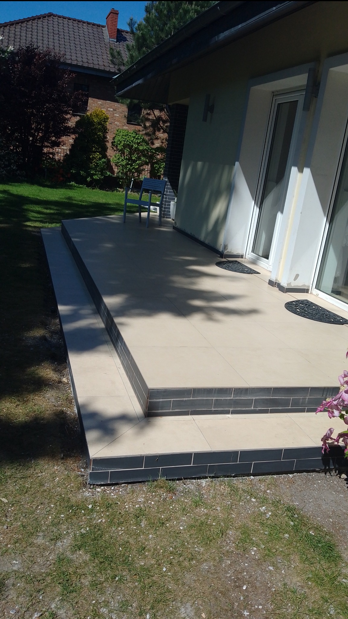 Wyłożony jasnymi płytkami taras z dwoma stopniami, obłożonymi ciemną glazurą, przylegający do budynku z białymi oknami i drzwiami, na zewnątrz widoczny trawnik i krzewy.