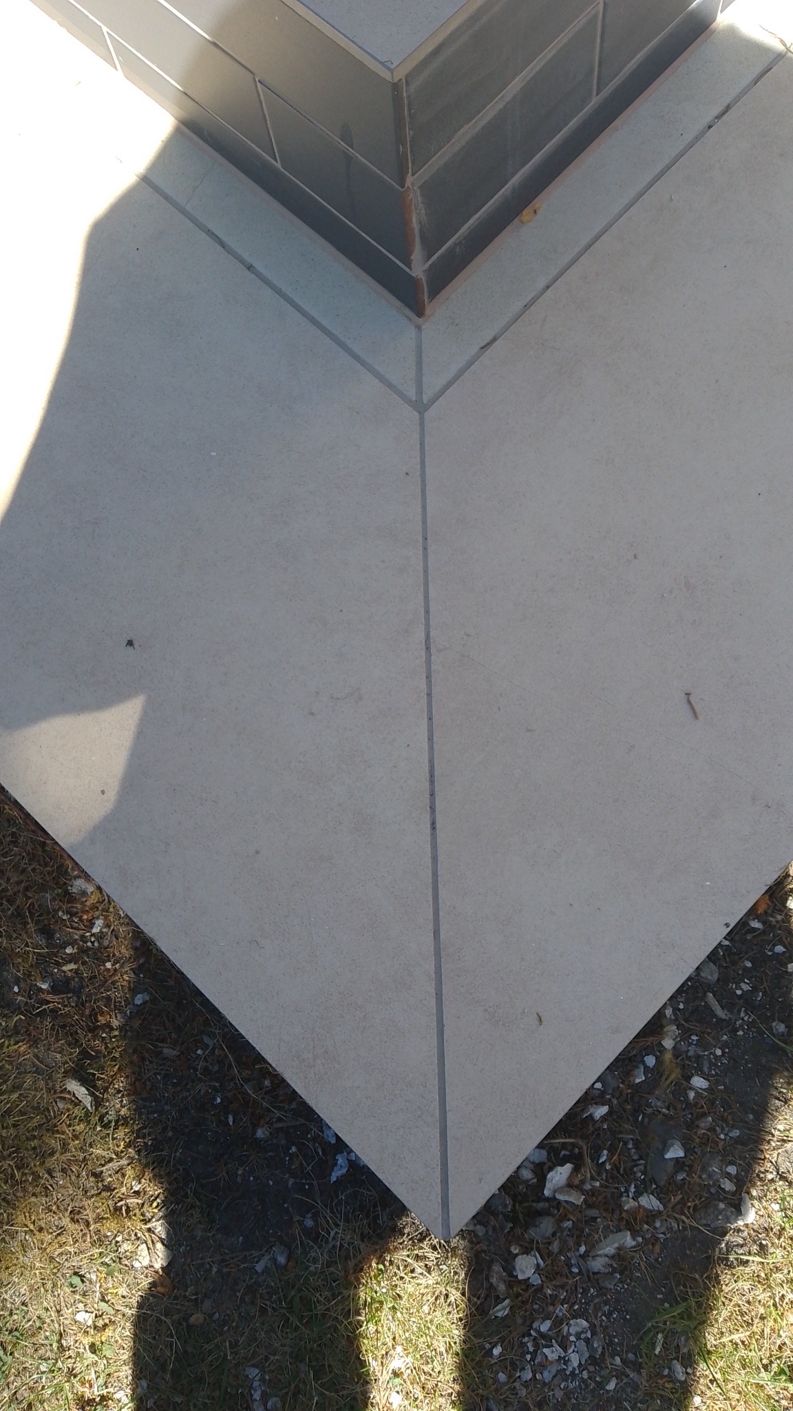 Szare płytki podłogowe ułożone na zewnątrz, z widocznymi fugami i fragmentem ściany wykończonej ciemnymi płytkami, oraz fragmentem trawy i ziemi.