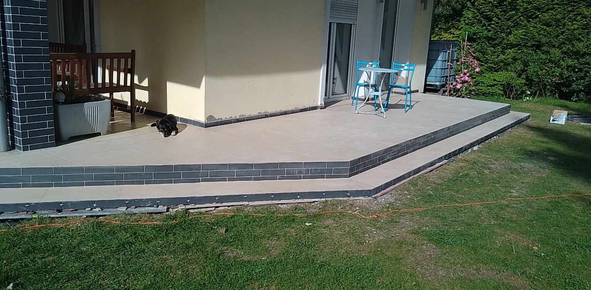 Nowo wyłożony, dwupoziomowy taras z szarych płytek z ciemnoszarym obramowaniem, z niebieskim stolikiem i krzesłami, oraz czarnym psem leżącym na górnym poziomie, w tle zielony ogród.