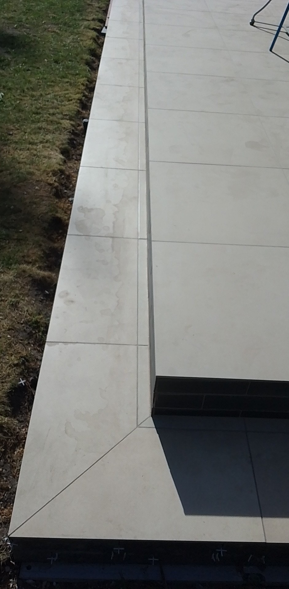 Ułożona, jasnoszara, ceramiczna podłoga tarasu z widocznymi fugami i plamami, z krawędzią wykończoną ciemniejszym materiałem, przylegająca do trawnika.