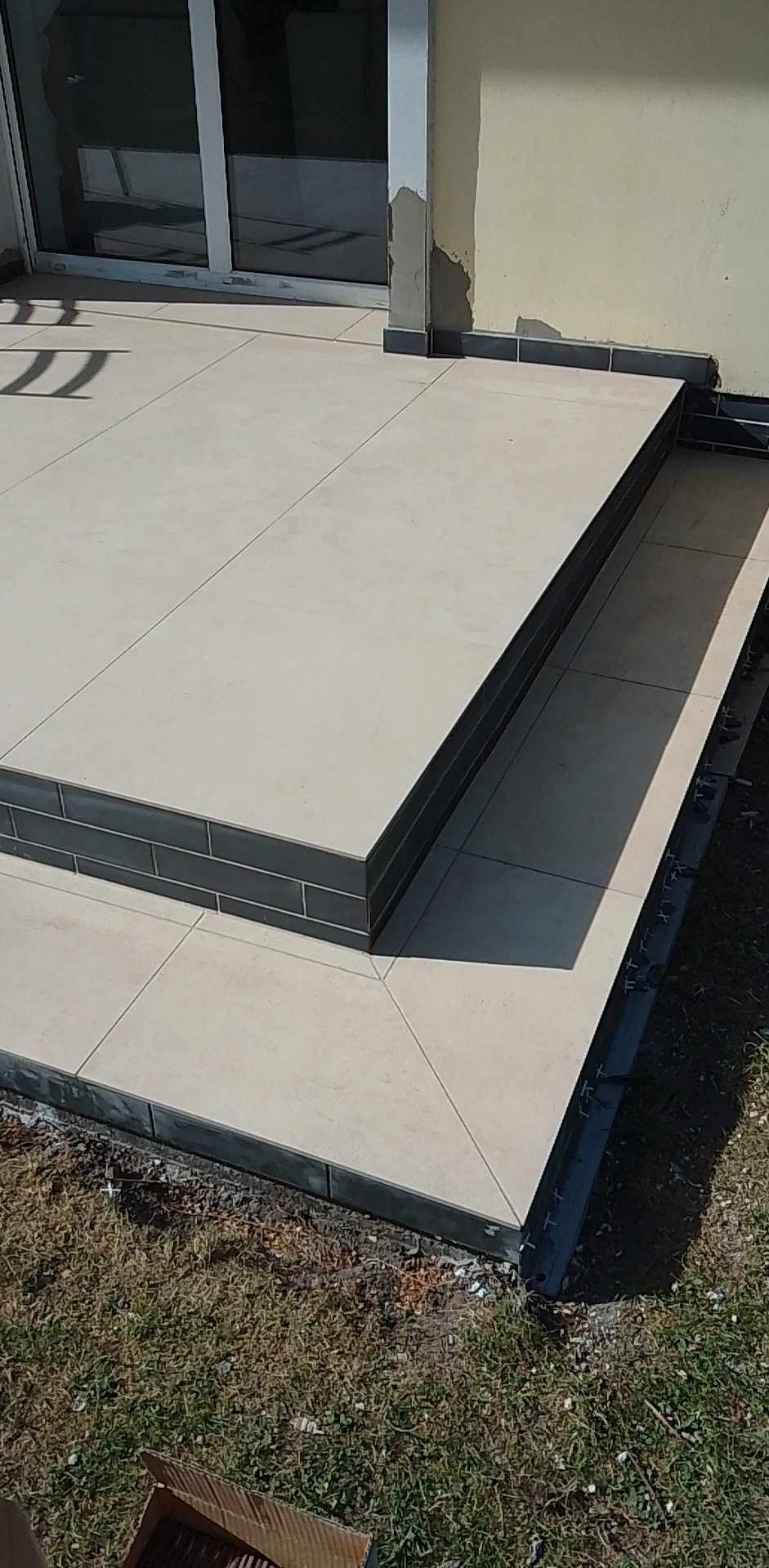 Wykończone schody zewnętrzne z jasnych płytek ceramicznych z ciemnoszarą krawędzią, widoczne elementy mocujące płytki na trawie.