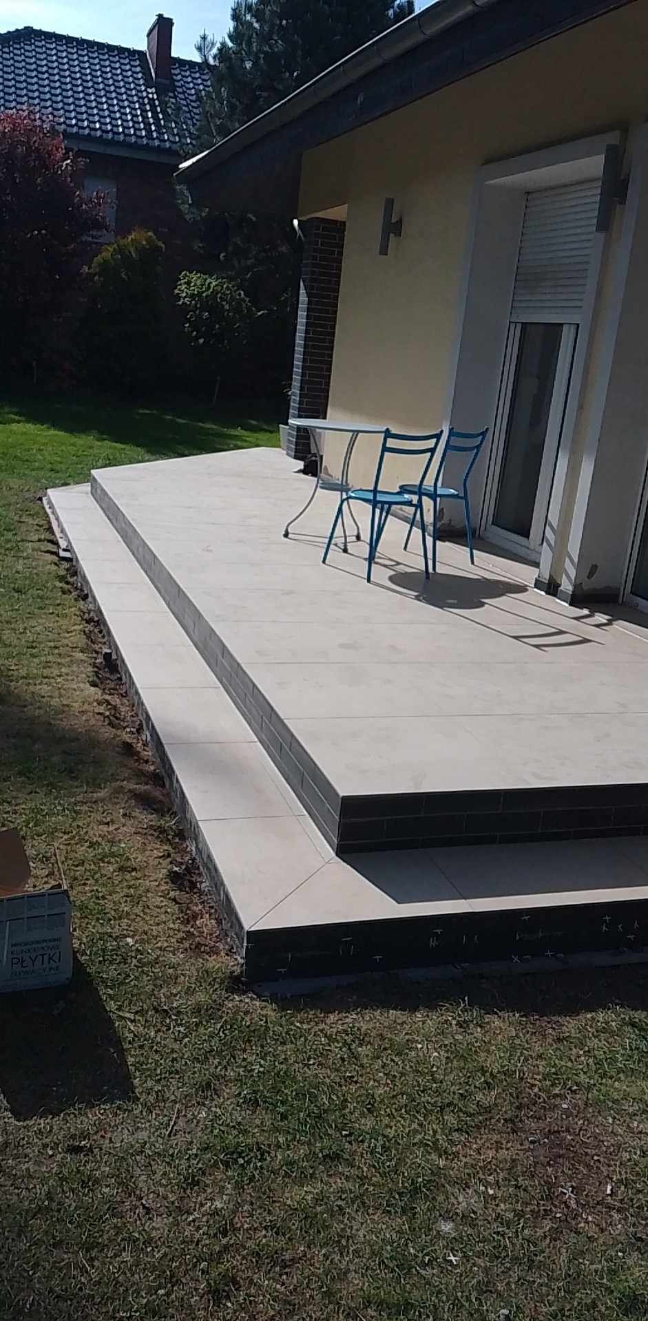 Wykończony, dwupoziomowy taras z jasnoszarych płytek z ciemnoszarymi obrzeżami, na którym stoją dwa niebieskie krzesła i okrągły stolik, widok z poziomu trawnika.