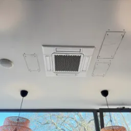 Biała, sufitowa klimatyzacja kasetonowa w jasnym pomieszczeniu. Widoczne ślady po montażu, lampy i okno z widokiem na drzewo.