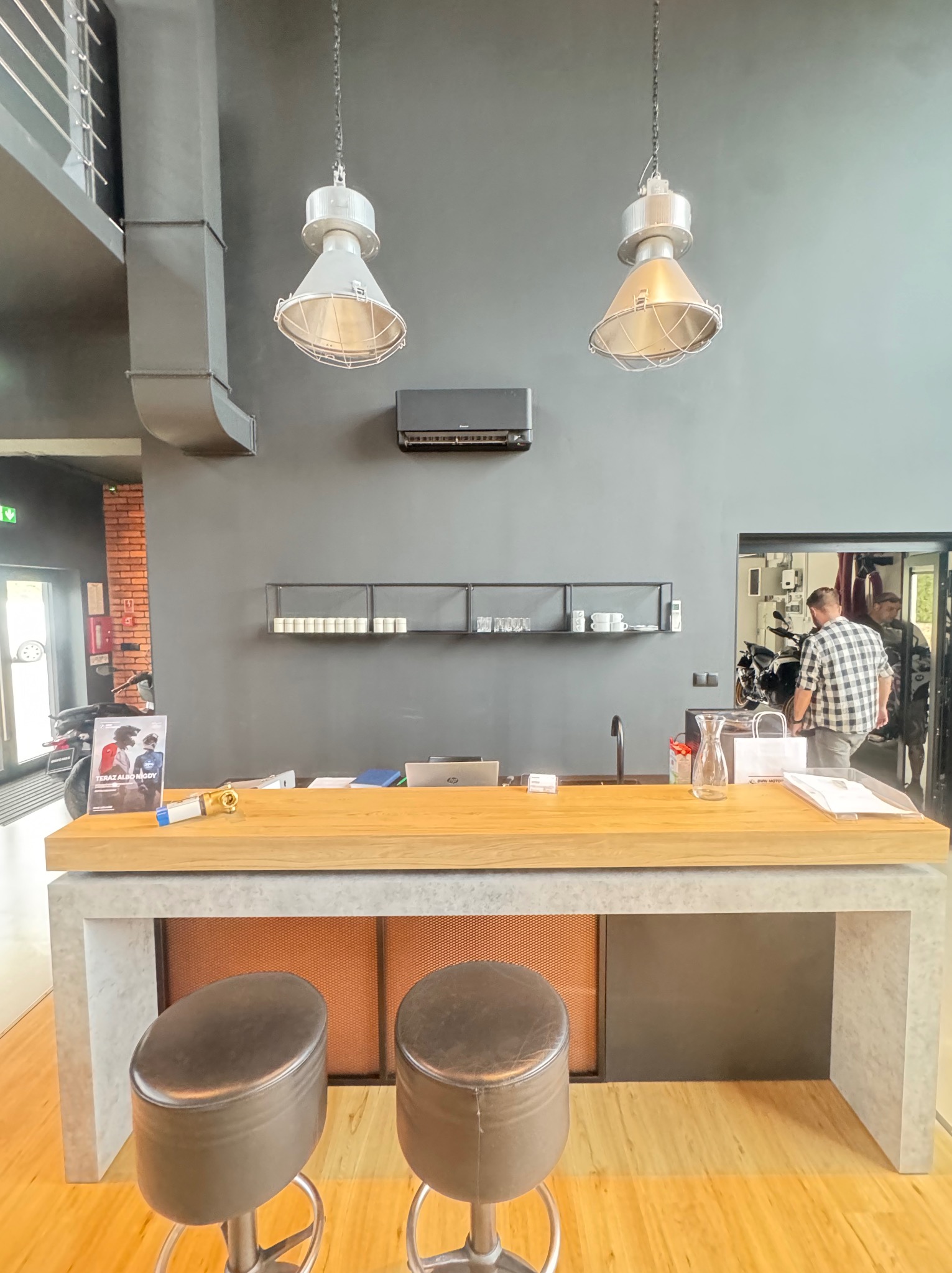 Nowoczesne wnętrze z klimatyzacją: szara ściana, industrialne lampy wiszące nad drewnianym blatem i hokerami. Widoczny klimatyzator i wentylacja.