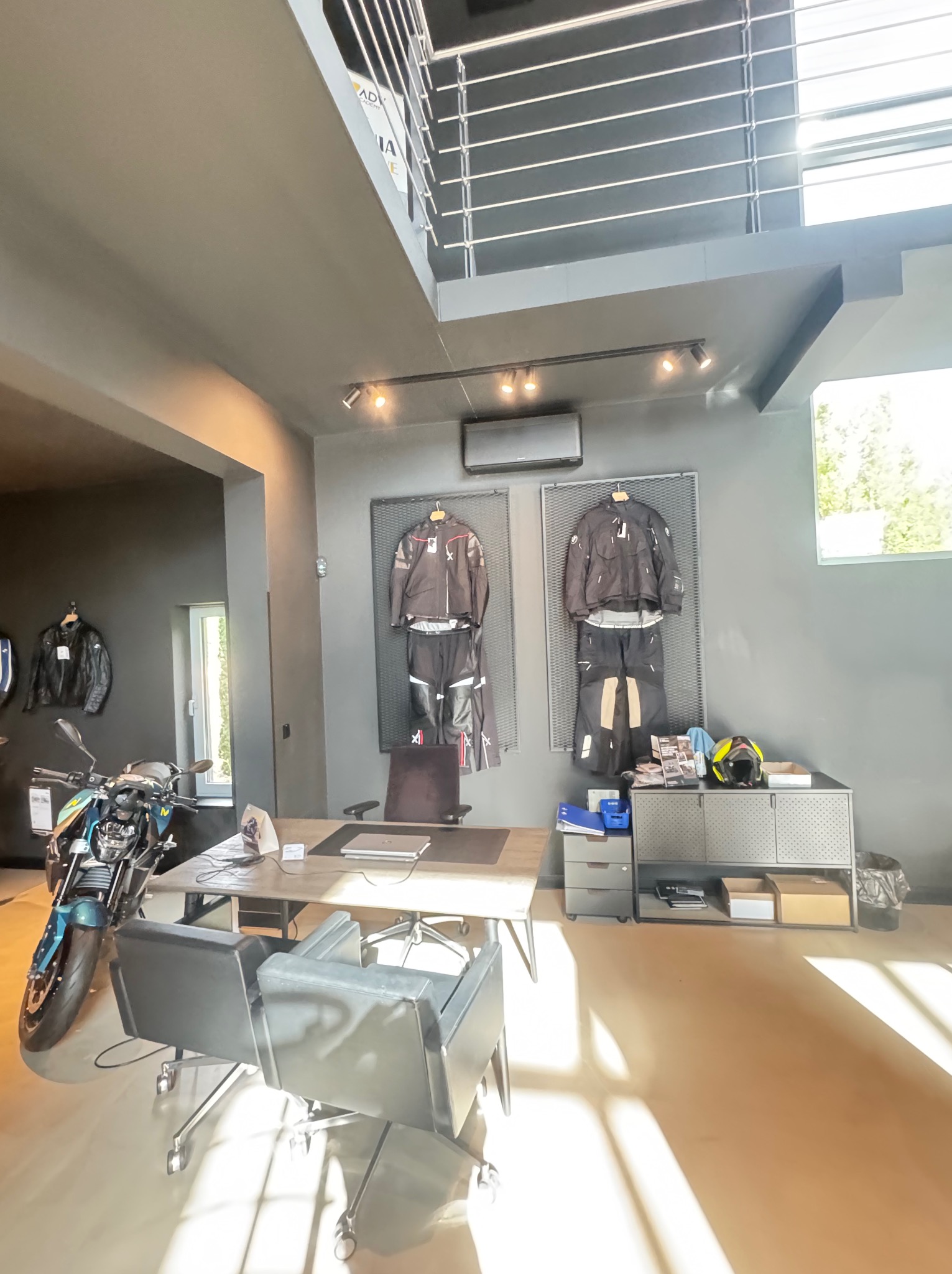 Nowoczesne biuro z klimatyzacją: motocykl, biurko, i ubrania motocyklowe. Klimatyzator dyskretnie zamontowany pod sufitem, oświetlenie punktowe.