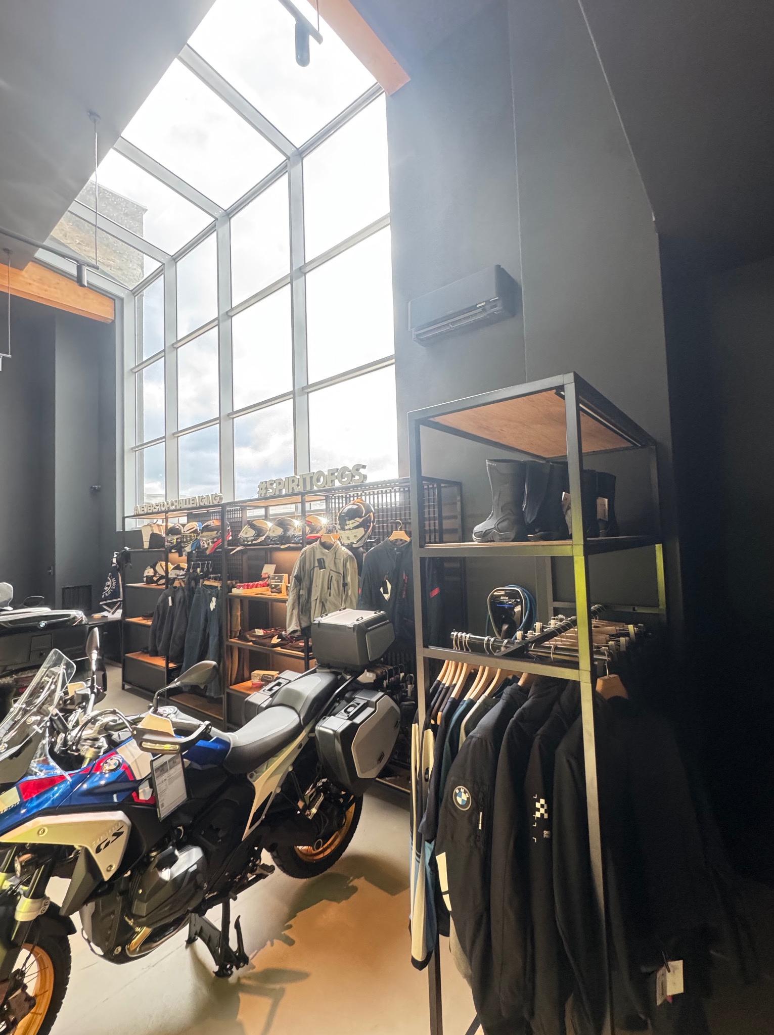 Salon motocyklowy z klimatyzacją: motocykle BMW, odzież motocyklowa na wieszakach i klimatyzator na ścianie. Duże okno dachowe w tle.