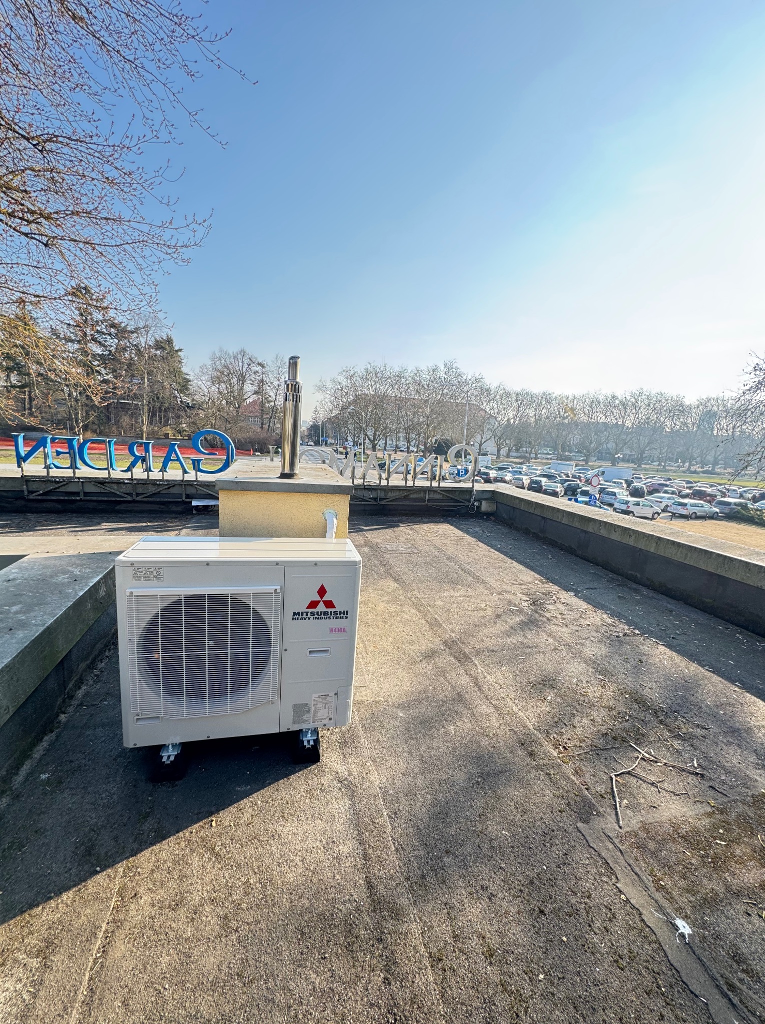 Biała jednostka zewnętrzna klimatyzacji Mitsubishi na dachu z widokiem na parking i drzewa w słoneczny dzień. Urządzenie na kółkach, z kominem wentylacyjnym obok.