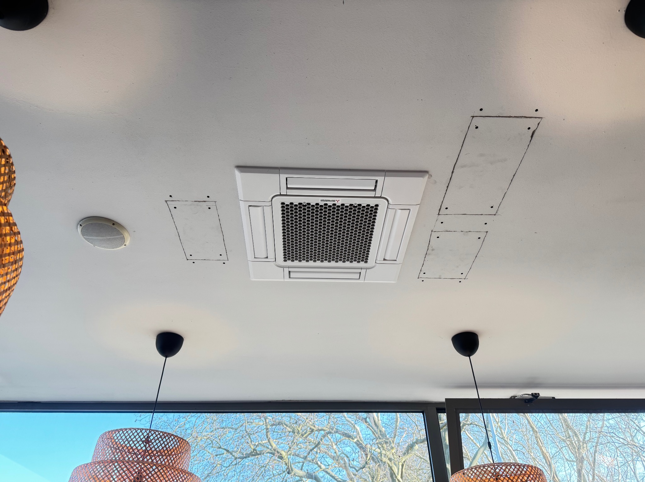 Biała, sufitowa klimatyzacja kasetonowa w jasnym pomieszczeniu. Widoczne ślady po montażu, lampy i okno z widokiem na drzewo.