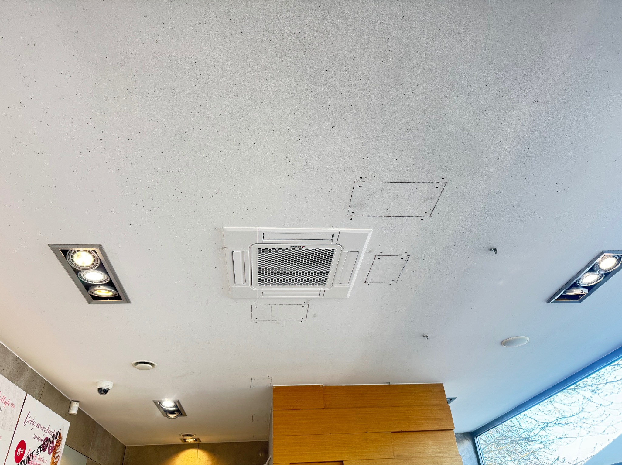 Kasetonowa klimatyzacja sufitowa w lokalu komercyjnym, widok z dołu. Biała obudowa, kratka wentylacyjna, panele rewizyjne. Oświetlenie punktowe i okno z widokiem na drzewo.