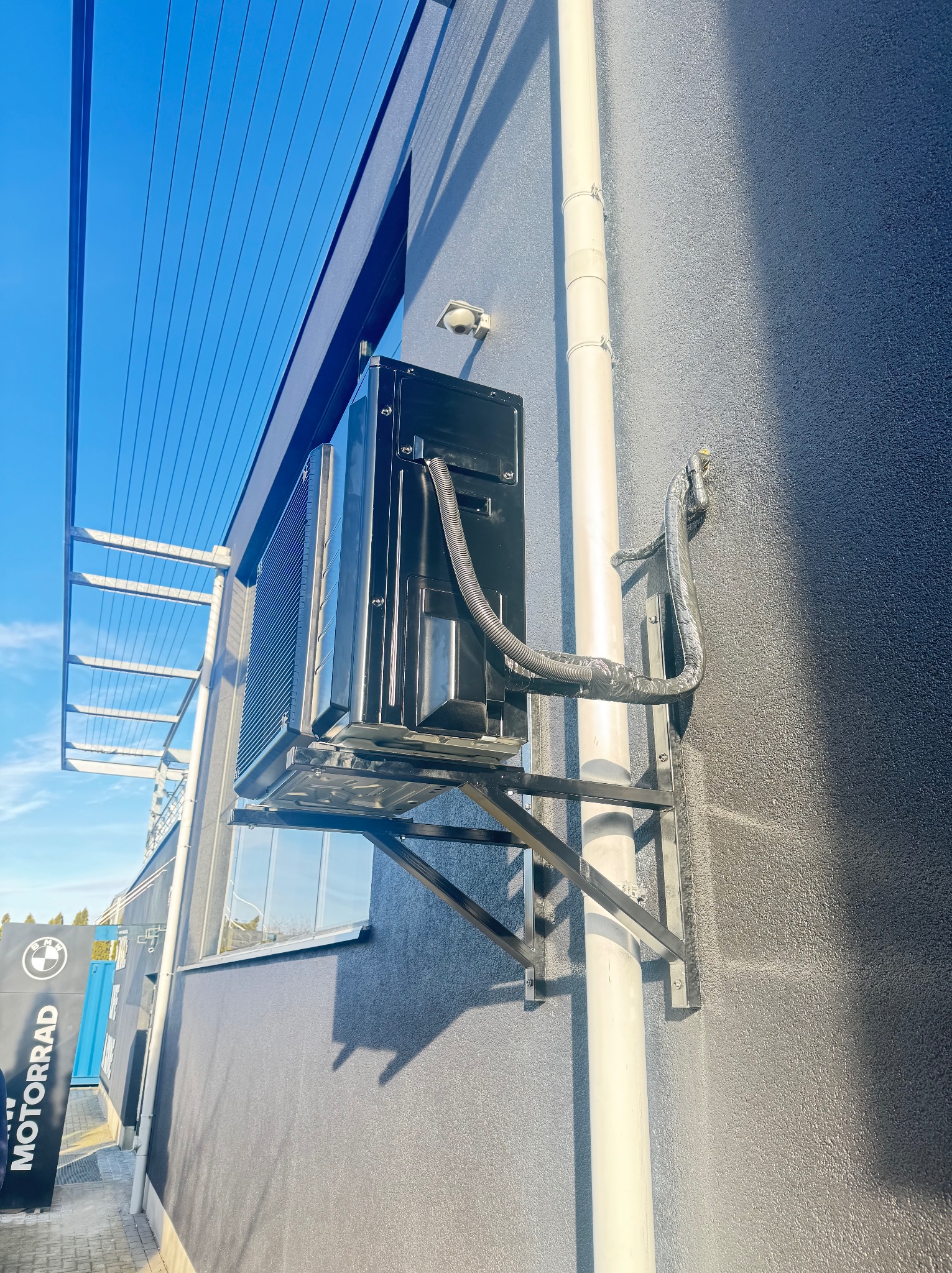 Czarna klimatyzacja zamontowana na szarej ścianie budynku, widoczne rury i wsporniki, z boku logo MOTORRAD, jasne niebo w tle.