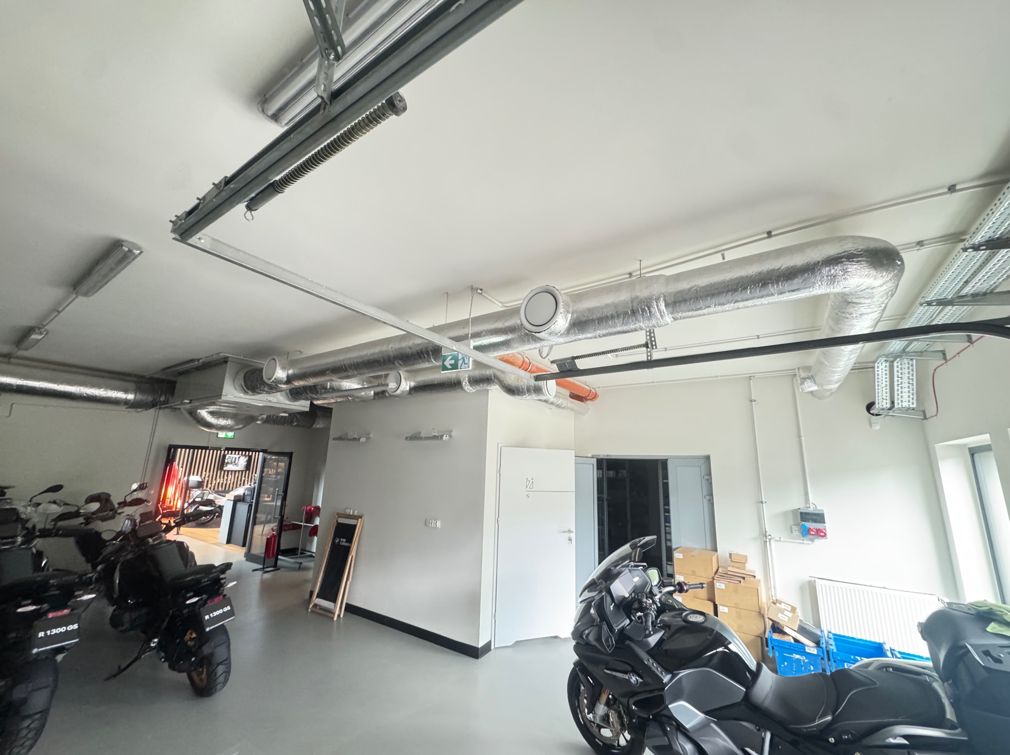 Instalacja wentylacji mechanicznej w lokalu w Szczecinie. Widoczne kanały wentylacyjne pod sufitem w pomieszczeniu z motocyklami. Srebrne rury i białe nawiewniki.