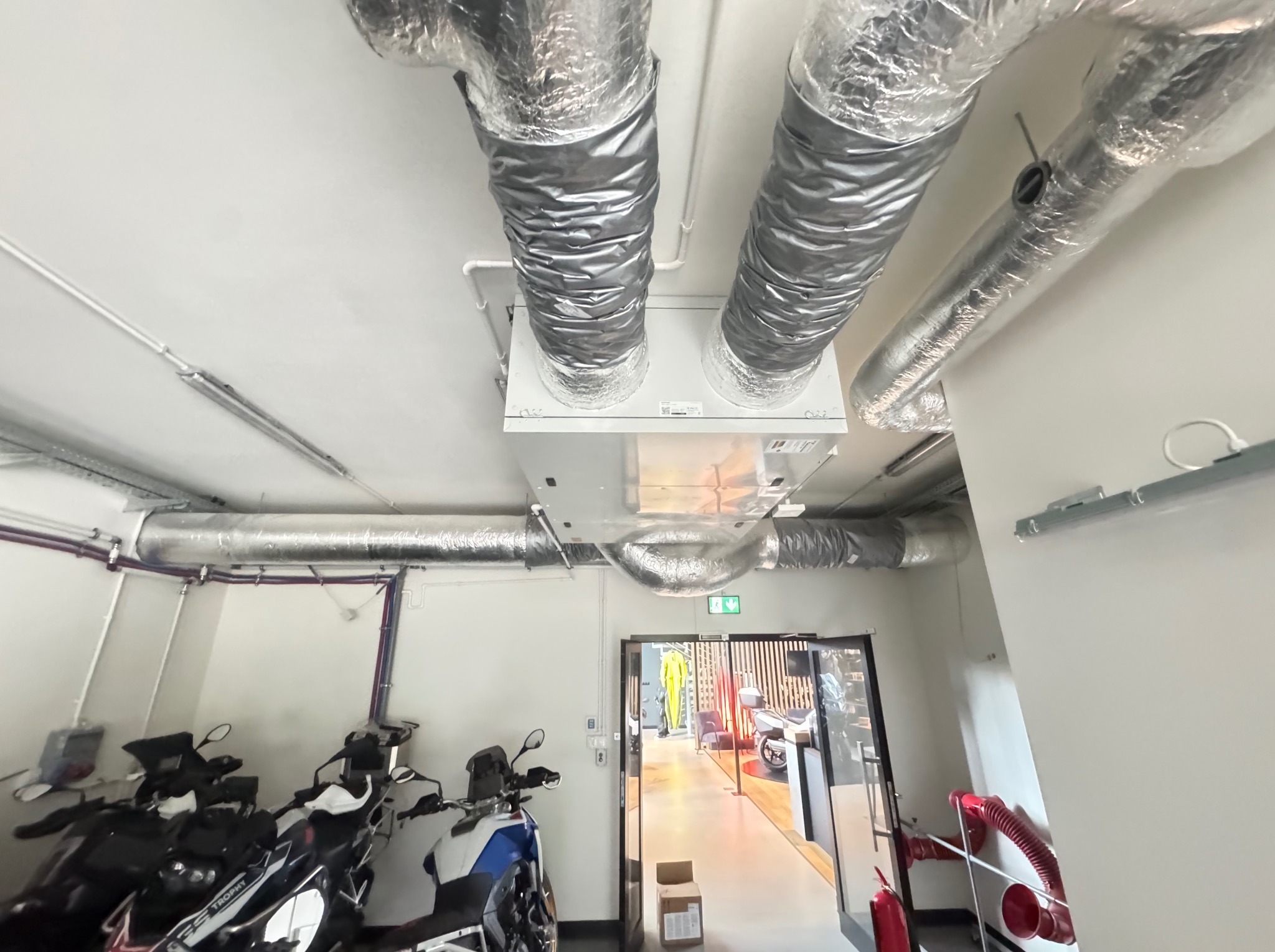 Biała centrala wentylacyjna z aluminiowymi kanałami w garażu z motocyklami. Widok z dołu, oświetlenie sufitowe, otwarte drzwi do salonu.