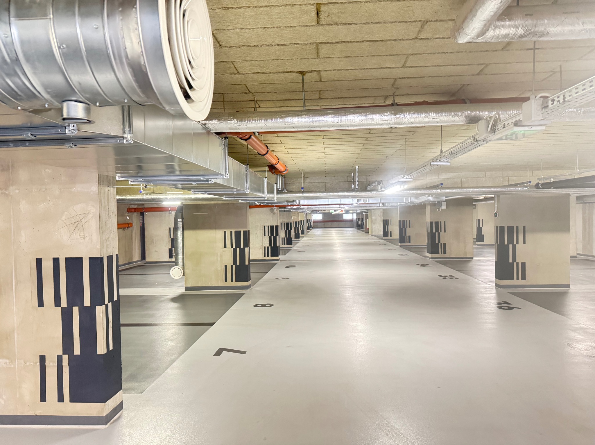 Instalacja wentylacyjna w garażu podziemnym: widoczne kanały wentylacyjne pod sufitem, rury i elementy mocujące. Perspektywa długa, z numeracją miejsc parkingowych.