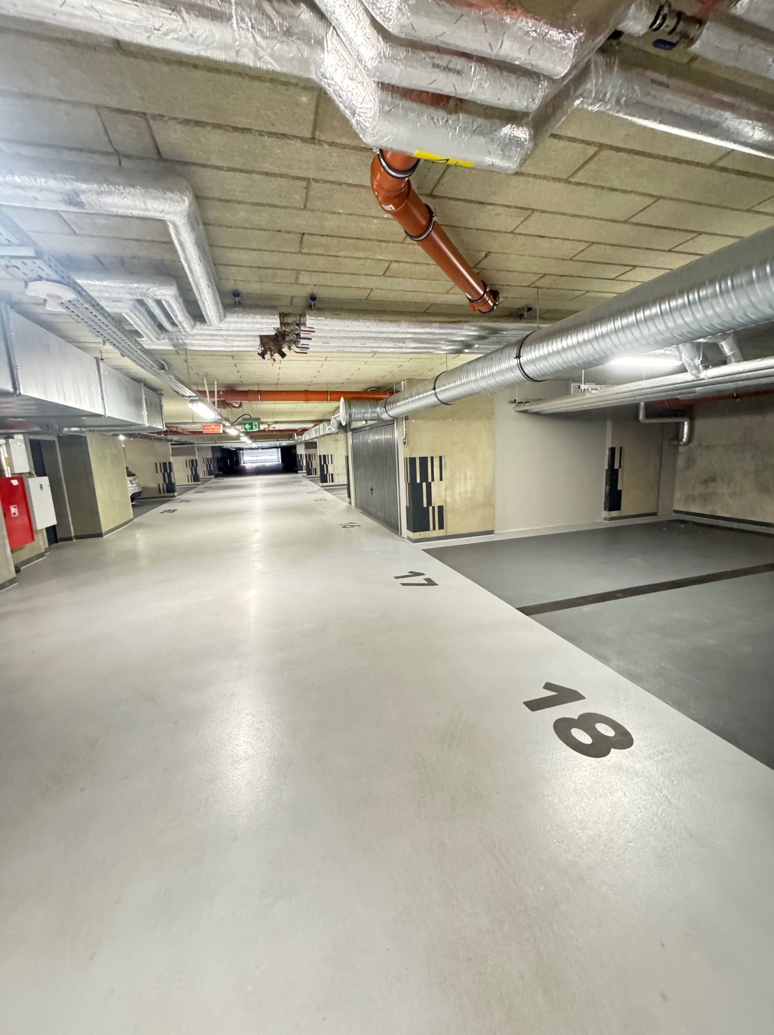 Instalacje wentylacyjne i rury w garażu podziemnym. Widoczne przewody w izolacji, rury wentylacyjne i instalacje pod stropem garażu z numerami miejsc parkingowych.