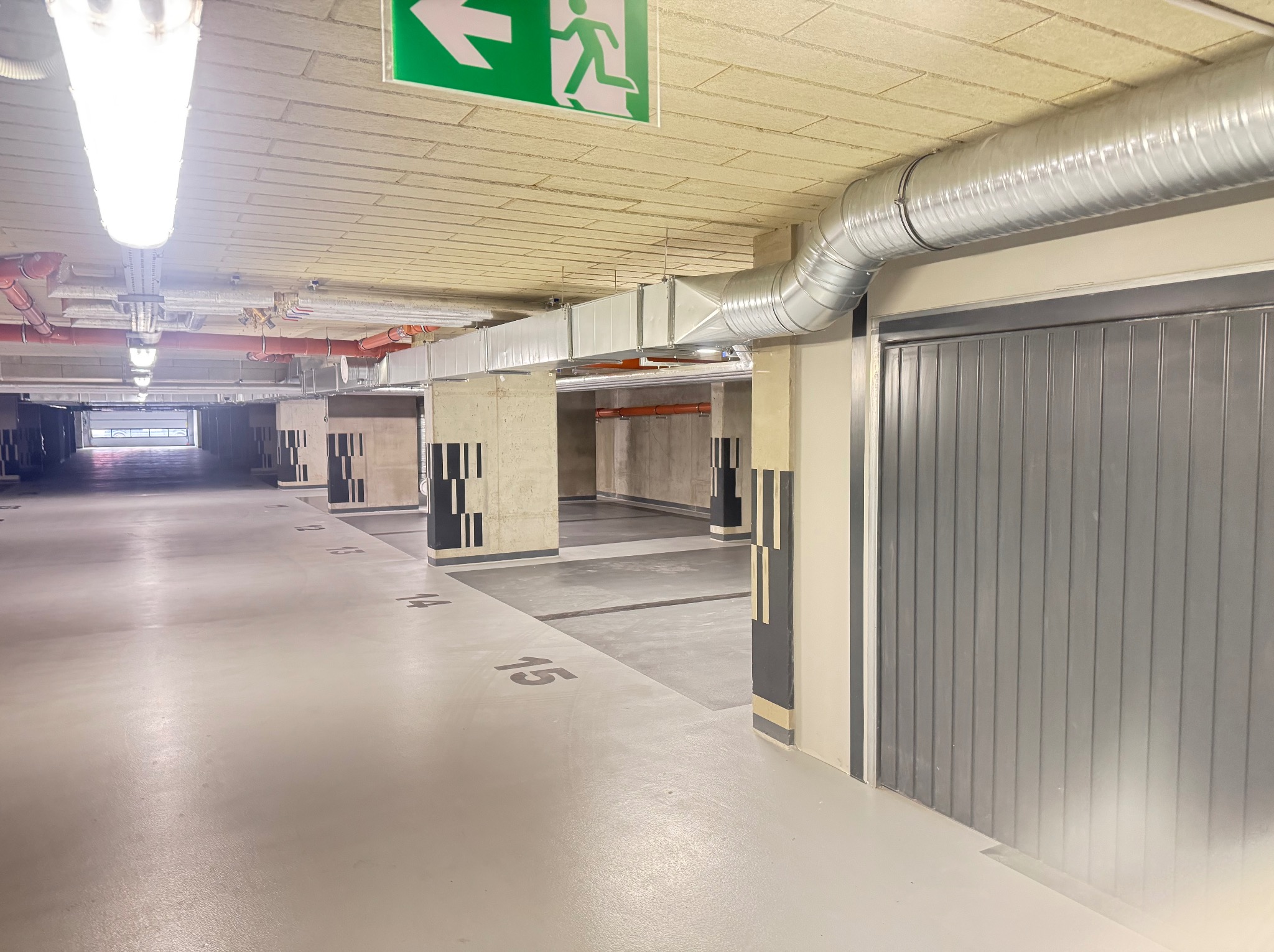 Instalacja wentylacyjna z rurami i kanałami w garażu podziemnym, widoczny znak ewakuacyjny. Szara brama garażowa na pierwszym planie, numeracja miejsc parkingowych na posadzce.