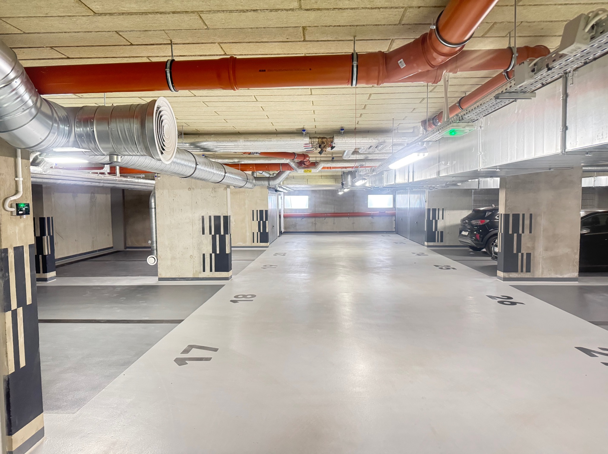 Instalacja wentylacyjna i rury w garażu podziemnym. Widoczne numery miejsc parkingowych na jasnej posadzce i betonowe filary z dekoracyjnymi pasami.